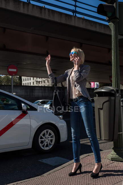 Jovem mulher de negócios elegante gesticulando para parada de táxi e falando no smartphone na rua da cidade — Fotografia de Stock