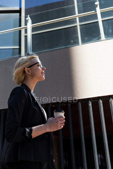 Seitenansicht der fröhlichen blonden Geschäftsfrau in stylischer Jacke mit Pappbecher an windigen Tagen — Stockfoto