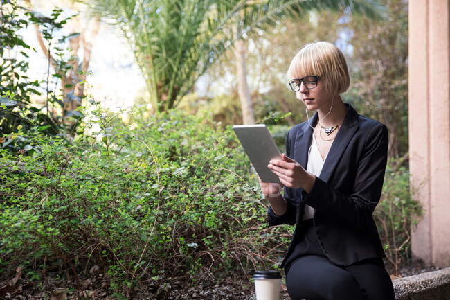 Молода блондинка, що сидить з паперовою чашечкою і користується цифровим планшетом в саду. — стокове фото