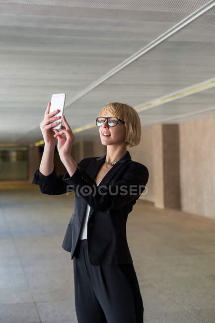 Білявка бізнес-леді в формальному одязі приймає селфі або перегляд смартфона у великому залі — стокове фото
