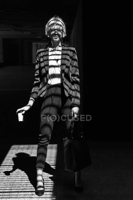 Чорно-білий знімок стильної жінки в офіційному одязі, що стоїть біля вікна з паперовою чашкою і дивиться на камеру — стокове фото