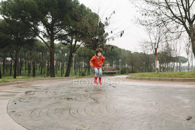 Чарівна радісна дитина в червоному плащі і гумових черевиках весело стрибає в калюжу на вулиці в парку в сірий день — стокове фото