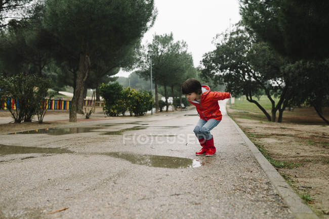 Criança alegre adorável em capa de chuva vermelha e botas de borracha se divertindo pulando na poça na rua no parque em dia cinza — Fotografia de Stock