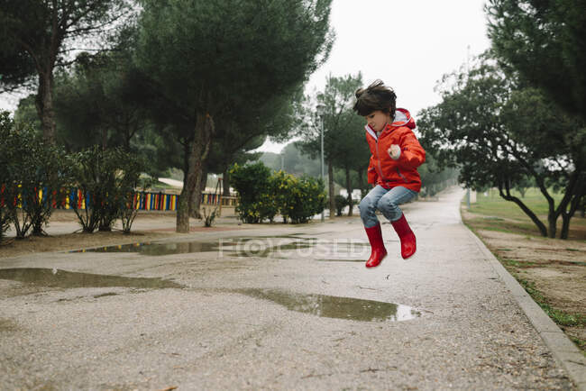 Criança alegre adorável em capa de chuva vermelha e botas de borracha se divertindo pulando na poça na rua no parque em dia cinza — Fotografia de Stock