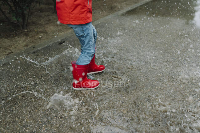 Adorable joyful child in red raincoat and rubber boots having fun jumping in puddle on street in park in gray day — Stock Photo