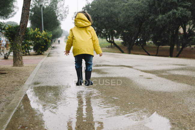 Back view of anonymous adorable joyful child in yellow raincoat and rubber boots having fun playing jumping in puddle on street in park in gray day — Stock Photo