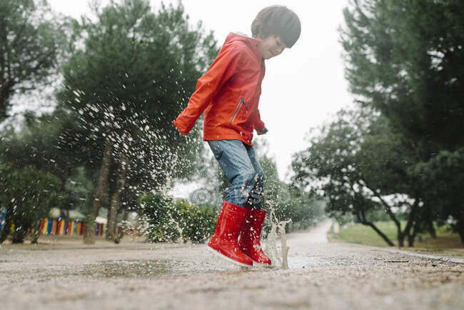 Adorable niño alegre en impermeable rojo y botas de goma que se divierten saltando en charco en la calle en el parque en día gris - foto de stock