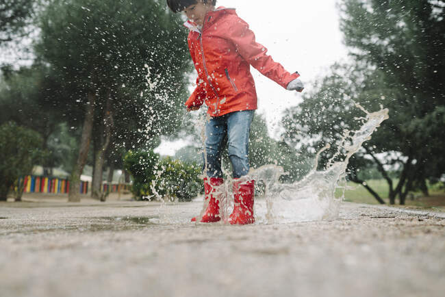 Adorable niño alegre en impermeable rojo y botas de goma que se divierten saltando en charco en la calle en el parque en día gris - foto de stock