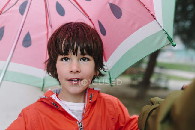 Aktives Kind mit Wassermelone im Stil öffnet Regenschirm in rotem Regenmantel und Gummistiefeln und blickt bei grauem Tag in die Kamera in der Parkgasse — Stockfoto