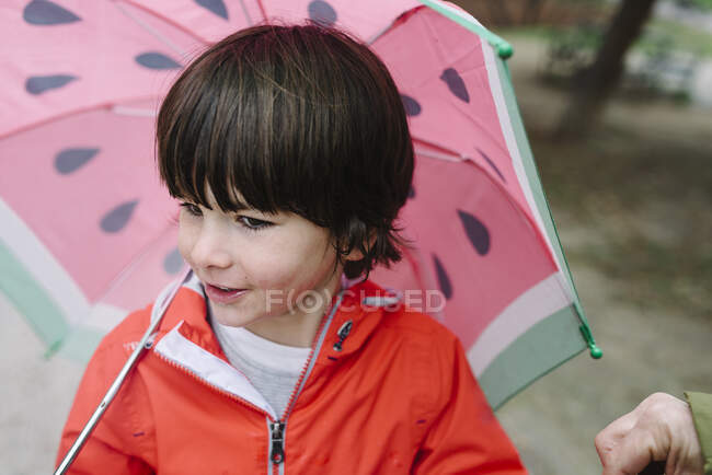 Активна дитина з кавуновими стилями відкриває парасольку в червоному плащі і гумових черевиках, дивлячись в сторону паркової алеї в сірий день — стокове фото