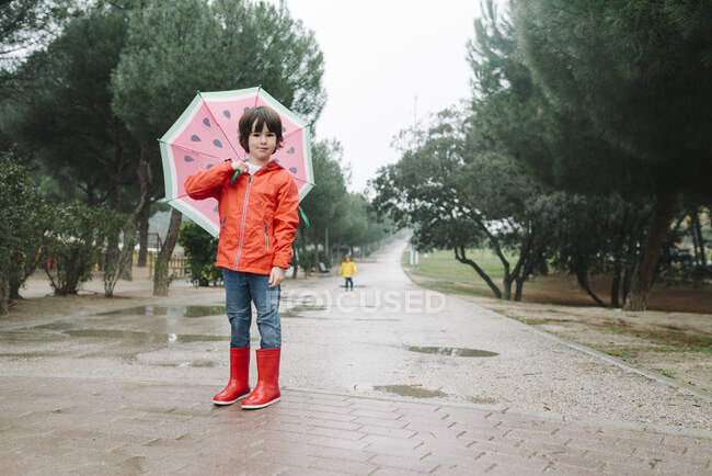 Active kid with watermelon styles open umbrella in red raincoat and rubber boots looking at camera in park alley in gray day — Stock Photo