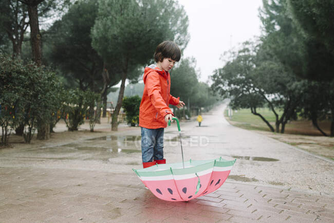 Active kid with watermelon styles open umbrella in red raincoat and rubber boots looking away in park alley in gray day — Stock Photo