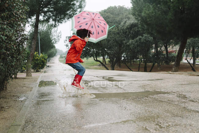 Seitenansicht aktives Kind mit Wassermelone Stile offenen Regenschirm in rotem Regenmantel und Gummistiefel springen in Park Gasse in grauen Tag — Stockfoto