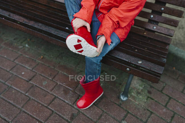 De cima cortado criança molhada irreconhecível em capa de chuva vermelha e botas de borracha sentado no banco de madeira e adiando a bota no parque em dia chuvoso — Fotografia de Stock
