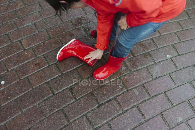 Curios kid pouring rain water from red rubber boot in wet park near wooden bench in gray day — Stock Photo