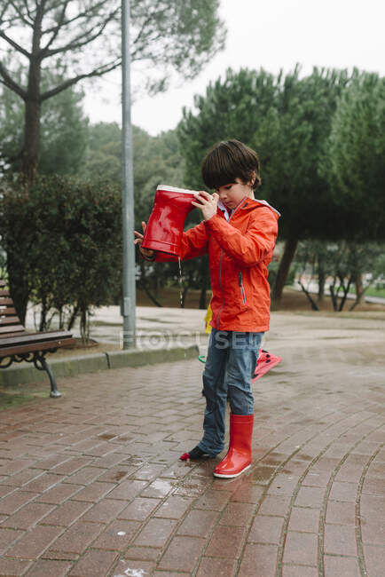 Curios bambino versando acqua piovana dallo stivale in gomma rossa nel parco bagnato vicino alla panchina in legno nella giornata grigia — Foto stock