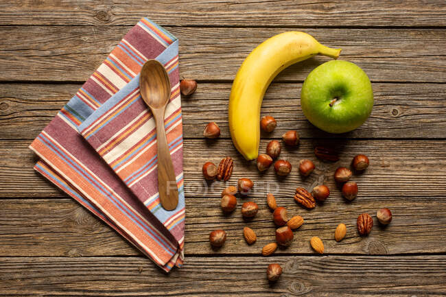 Вид сверху на яблоко и банан с орехами рядом с ложкой и игрушкой на деревянном столе со здоровой пищей — стоковое фото
