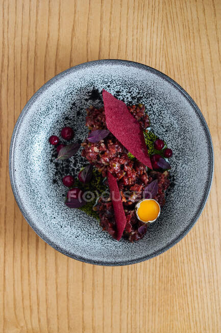 From above top view of ceramic bowl with salad made with marinated herring and wild red lingonberries garnished with raw quail egg on wooden table — Stock Photo
