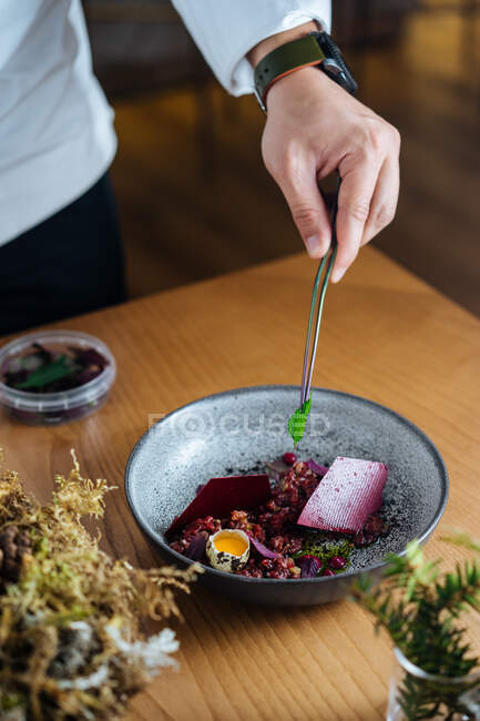 Zugeschnittene unkenntliche Person bei der Zubereitung auf einer Keramikschüssel mit Salat aus mariniertem Hering und wilden roten Preiselbeeren, garniert mit rohem Wachtelei auf dem Holztisch — Stockfoto