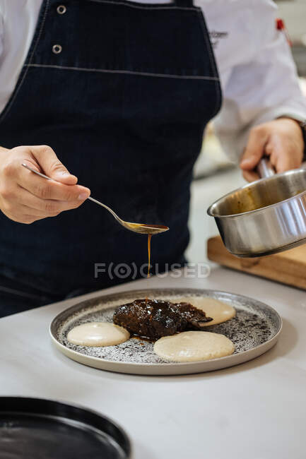 Crop chef restaurante com panela e colher em mãos derramando molho sobre a comida enquanto prepara o prato de cozinha haute elegante — Fotografia de Stock
