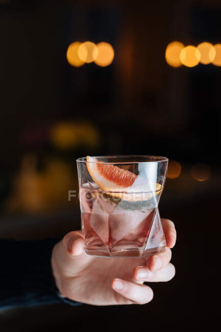 Ausgeschnittene unkenntliche Person hält Glas mit kaltem alkoholischen Cocktail mit Grapefruitscheibe und Eiswürfel auf dem Tisch vor schwarzem Hintergrund — Stockfoto