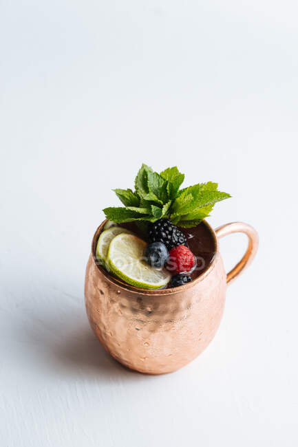Taza de metal de arriba con porción de sabrosa bebida de frutas con lima y bayas decoradas con hojas de menta y colocadas sobre fondo blanco - foto de stock
