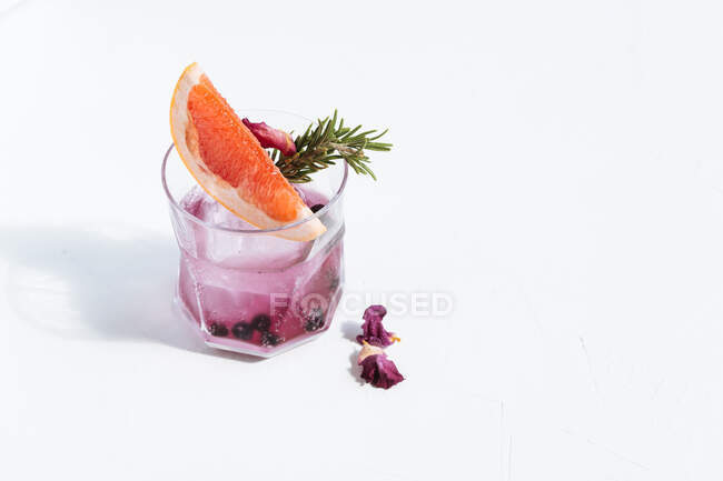 Copa con cóctel alcohólico frío con rodaja de pomelo, romero, arándano seco, flores y cubitos de hielo colocados sobre la mesa sobre fondo blanco - foto de stock