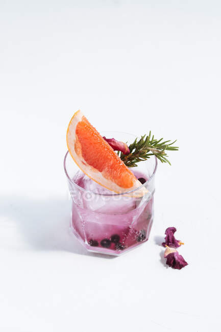 Glass with cold alcoholic cocktail with slice of grapefruit, rosemary, dry blueberry, flowers and ice cube placed on table against white background — Stock Photo