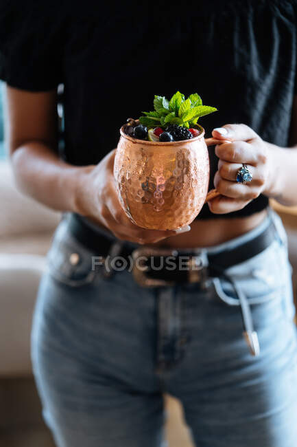 Неузнаваемая женщина несет металлическую кружку натурального фруктового напитка с ягодами и мятными листьями в летний день в кафе — стоковое фото