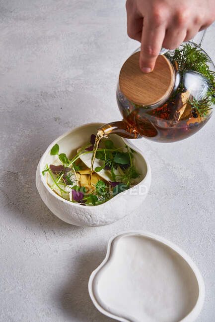 Personne méconnaissable verser du thé chaud frais de théière en verre dans un bol en céramique avec des herbes — Photo de stock