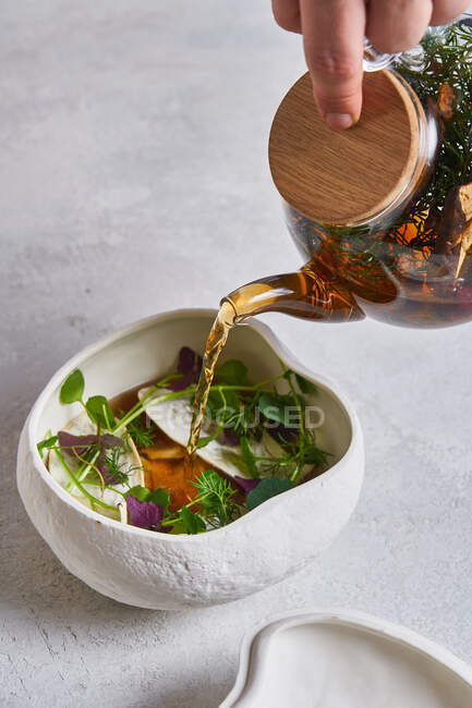 Unrecognizable person pouring fresh warm tea from glass teapot into ceramic bowl with herbs — Stock Photo