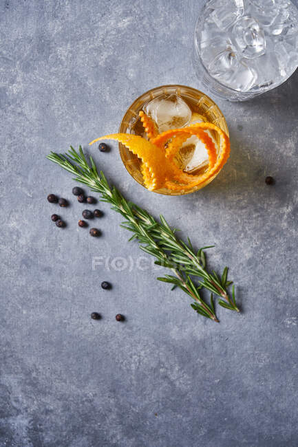 Зверху скляна чашка з холодним старомодним коктейлем з віскі та апельсиновою шкіркою, розміщена на сірому столі з розмариновою рослиною, перець зерна — стокове фото