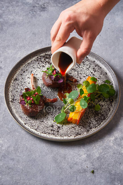 Von oben zugeschnittene unkenntliche Person, die schmackhafte Fleischmedaillons mit Kräutern auf Teller auf grauem Stucktisch mit Sauce würzt — Stockfoto