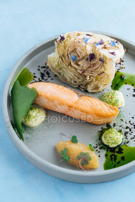 Du haut du filet de saumon cuit au four et du caviar de brochet avec un morceau de jeune chou sur une élégante assiette grise décorée de sauce blanche sur fond bleu — Photo de stock