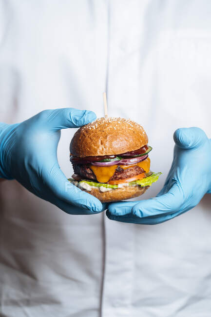 Unrecognizable cropped person hands with gloves holding burger with patty of venison fresh rye bread rolls red onion cheese slices of tomatoes lettuce leaf and sauce on plate with fried potato — Stock Photo