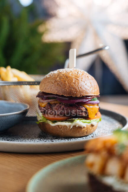 Appetizing burger with patty of venison fresh rye bread rolls red onion cheese slices of tomatoes lettuce leaf and sauce on plate with fried potato in restaurant — Stock Photo