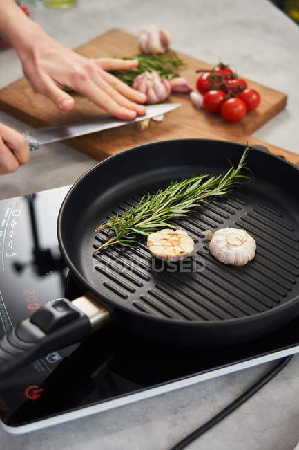 Grill pan with sprigs of fresh rosemary and cut garlic head placed on stove and crop hands of woman crushing garlic on wooden cut board in background — Stock Photo