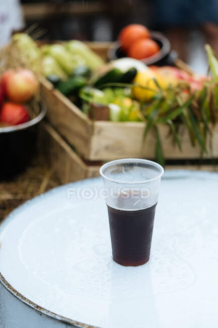 Сверху пластиковой чашки свежего тёмного пива возле коробки с различными овощами на рынке — стоковое фото