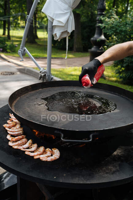 From above of crop chef in glove with bottle in hand pouring oil on pan near shrimps in marketplace — Stock Photo