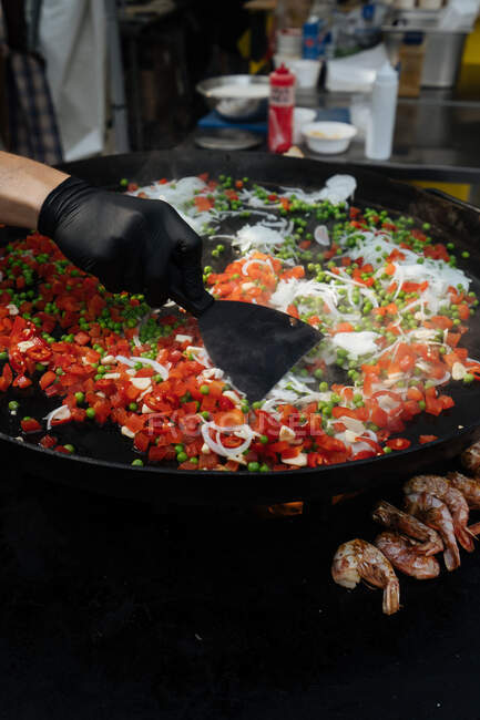 De cima do chef de colheita no prato de cozinha de luva de ervilha com tomate e cebola perto de camarões na panela de metal no mercado — Fotografia de Stock