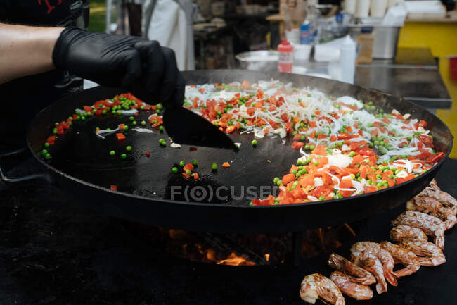From above of crop chef in glove cooking dish of pea with tomato and onion near shrimps on metal pan in marketplace — Stock Photo