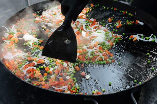 De cima do chef de colheita no prato de cozinha de luva de ervilha com tomate e cebola perto de camarões na panela de metal no mercado — Fotografia de Stock