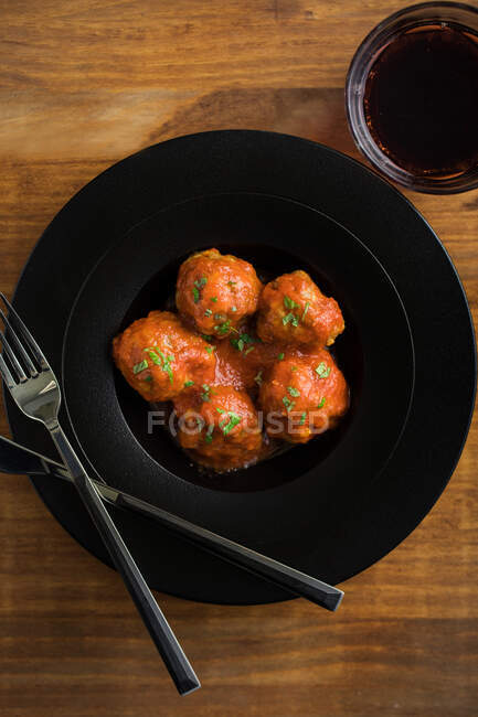 Сверху вкусные фрикадельки с томатным соусом, подаваемые с хлебом на черной тарелке со столовыми приборами и напитками на столе — стоковое фото