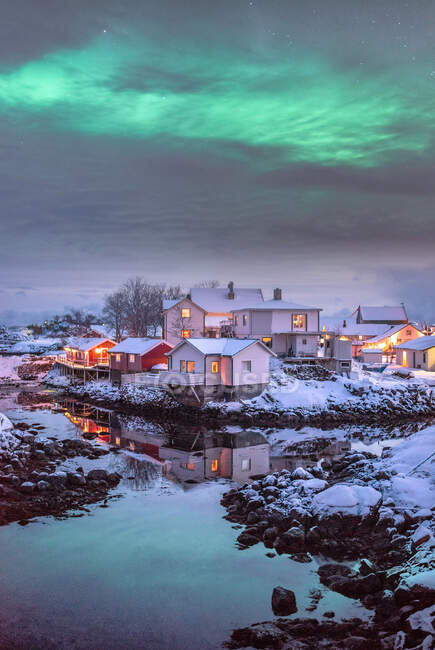 Des lumières polaires brillantes brillent au-dessus d'un joli petit village au bord d'une rivière isolée entourée de roches blanches et givrées en hiver à Svolvaer, en Norvège — Photo de stock