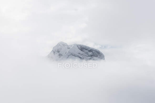 Rocky snowy mountain high in cloudy sky in foggy white day — Stock Photo