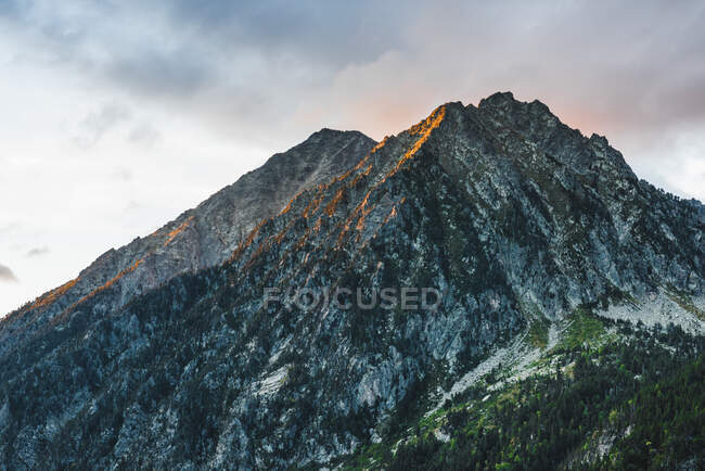 Величний природний пейзаж з скелястим піком гірського хребта, покритий зеленими рослинами на тлі хмарного неба з сонячним світлом — стокове фото