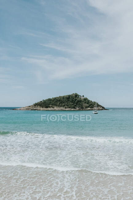 Пенная волна и бирюзовый океан, омывающий маленький каменистый остров с зелеными деревьями под облачным небом в яркий день — стоковое фото