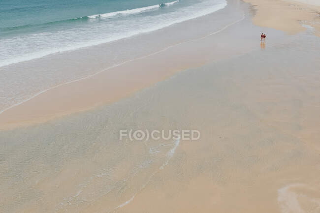 De arriba las ondas espumosas que lavan la costa arenosa con las personas que caminan en el día brillante - foto de stock