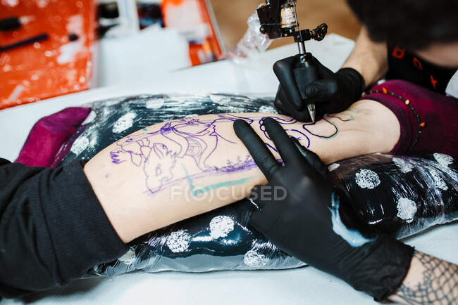 Dall'alto anonimo tatuatore ritagliato in guanti di lattice utilizzando la macchina del tatuaggio per fare schizzo sulla gamba del cliente del raccolto nel salone moderno — Foto stock