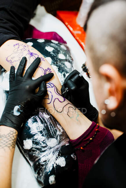 С вышесказанного анонимный мужчина-татуировщик в латексных перчатках с помощью тату-машины делает набросок на ноге клиента в современном салоне — стоковое фото